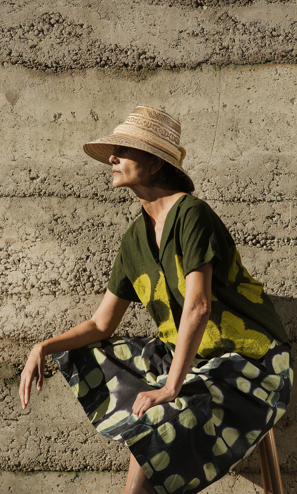 Plümo- A woman in a straw hat and Orla Flower Skirt sits against a textured concrete wall in sunlight.