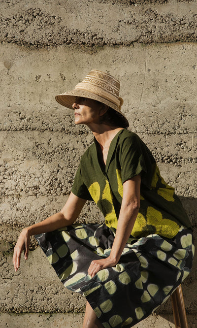 Plümo- A woman in a straw hat and Orla Flower Skirt sits against a textured concrete wall in sunlight.