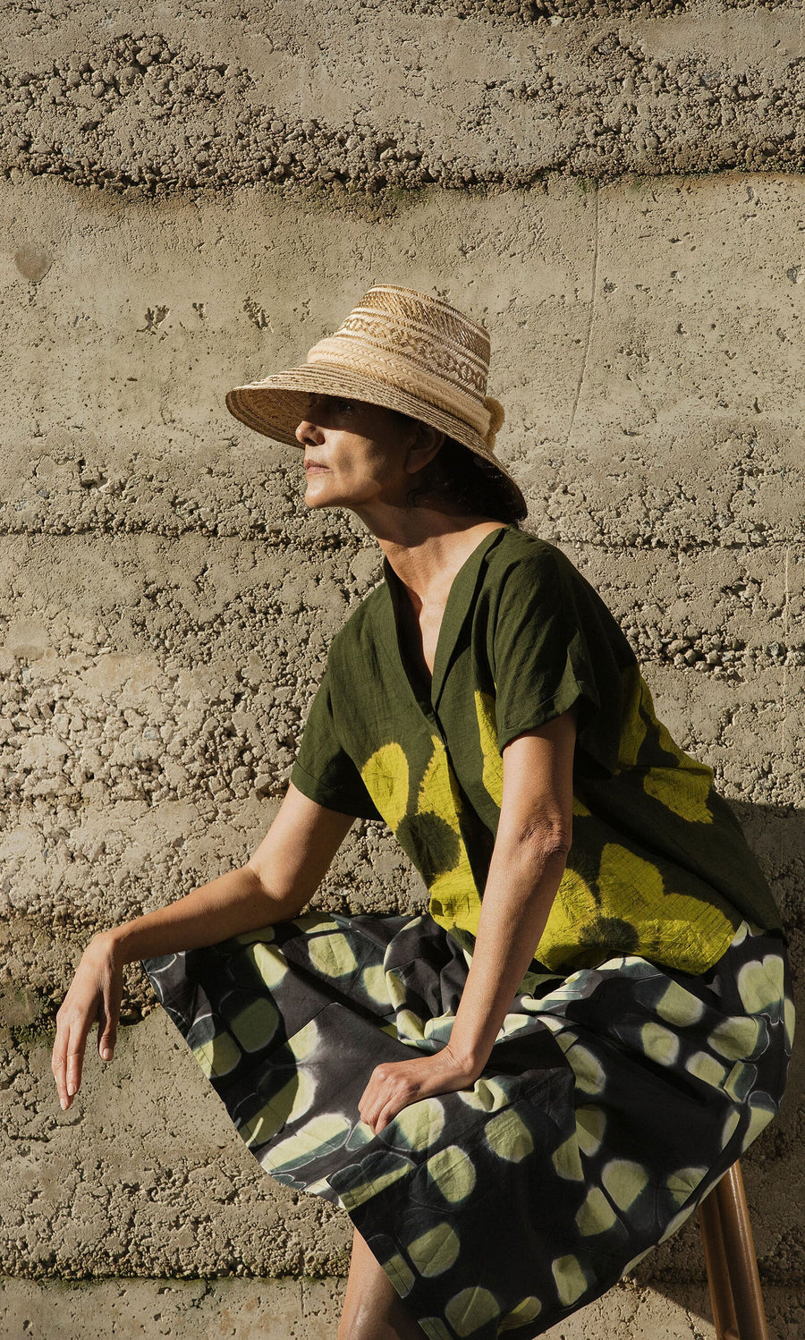 Plümo- A woman in a straw hat and Orla Flower Skirt sits against a textured concrete wall in sunlight.