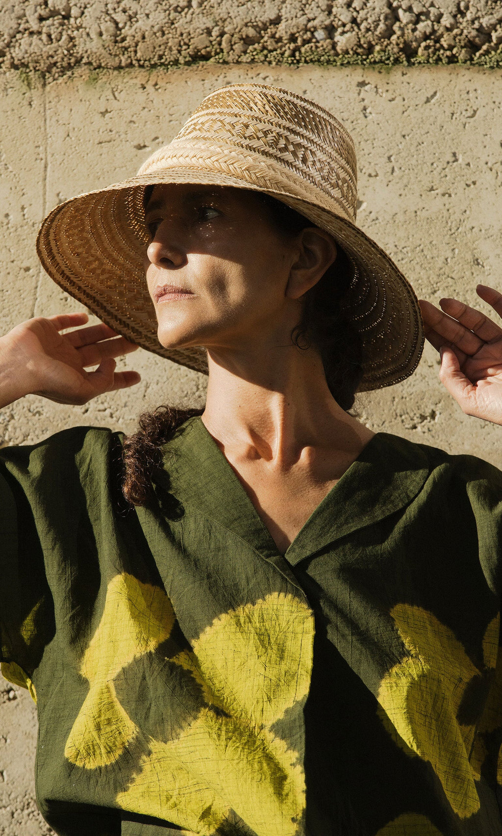 Plümo- Woman in a green shirt wears the Bogota Hat, standing by a textured wall in sunlight.