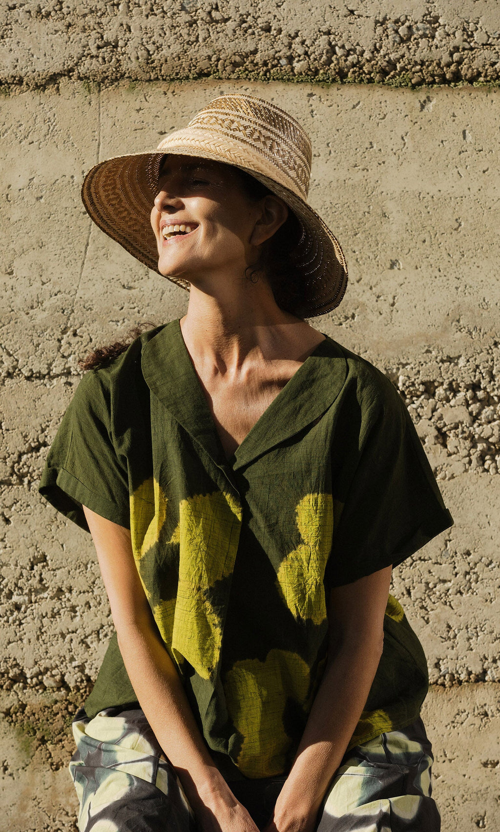 Plümo- A woman in an Orta Blouse and wide-brimmed hat smiles in sunlight against a textured wall.