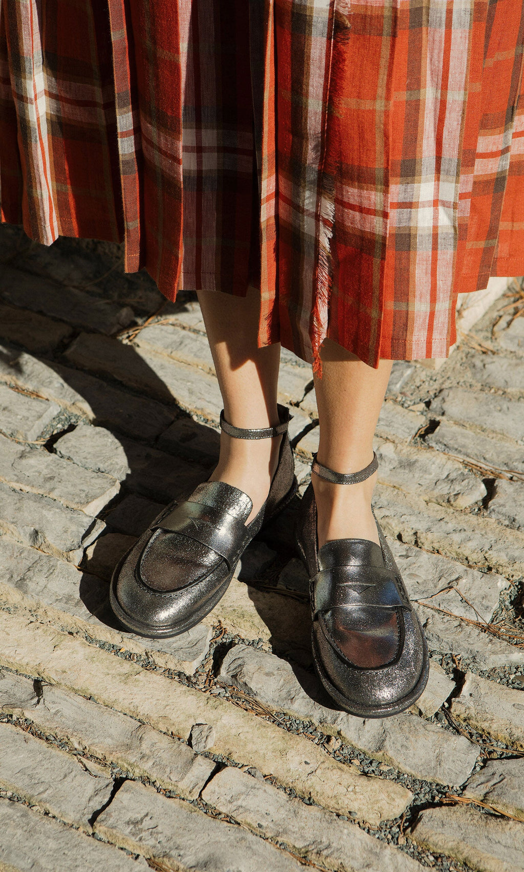 Plümo- Person wearing silver loafers and a red plaid skirt, standing on a cobblestone street in sunlight.