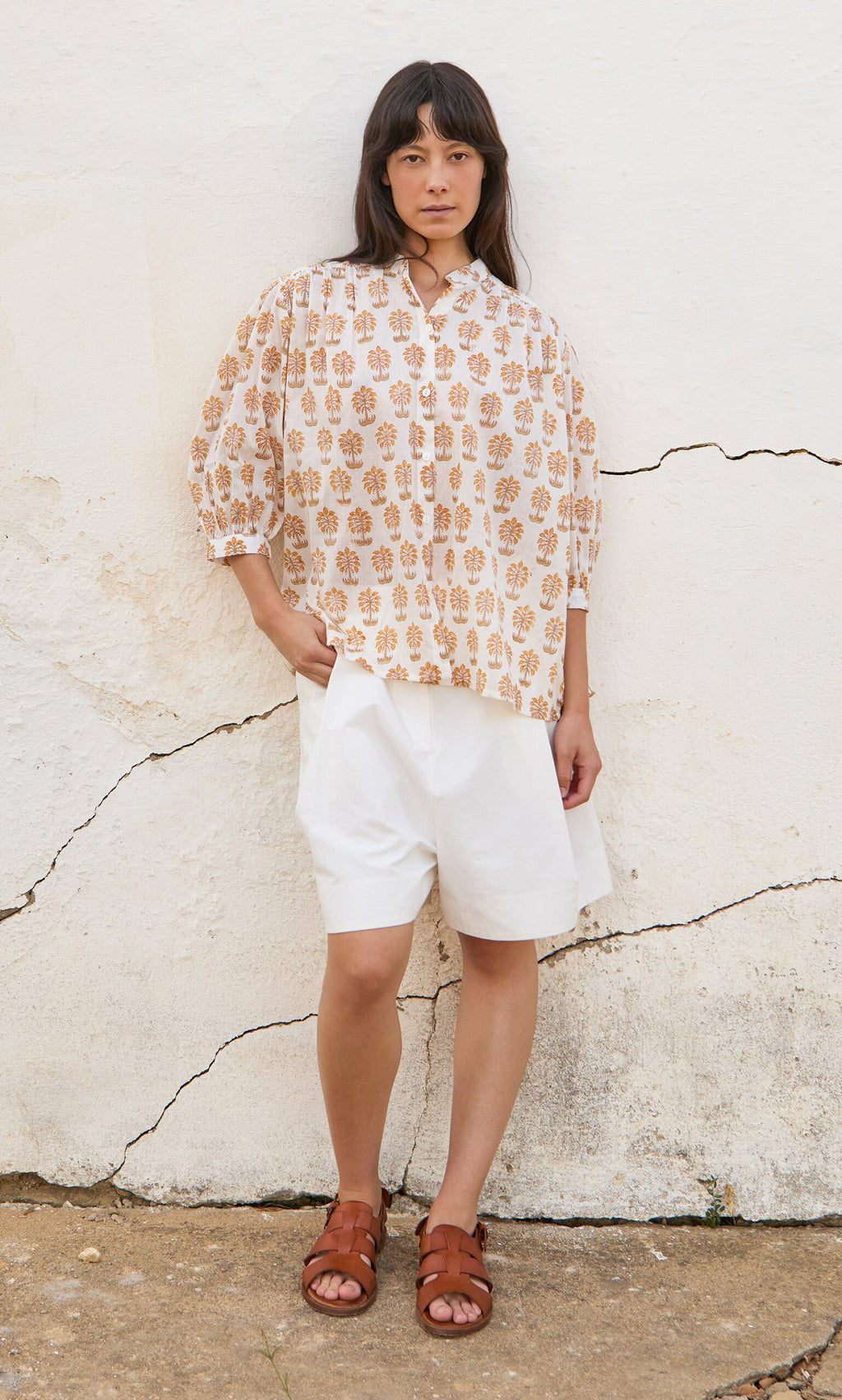 Plümo- Woman stands against a cracked white wall wearing the Palm Shirt, white shorts, and brown sandals.