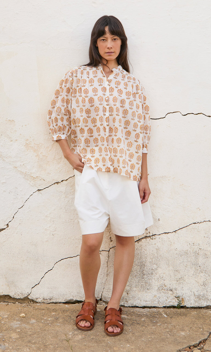 Plümo- Woman stands against a cracked white wall wearing the Palm Shirt, white shorts, and brown sandals.