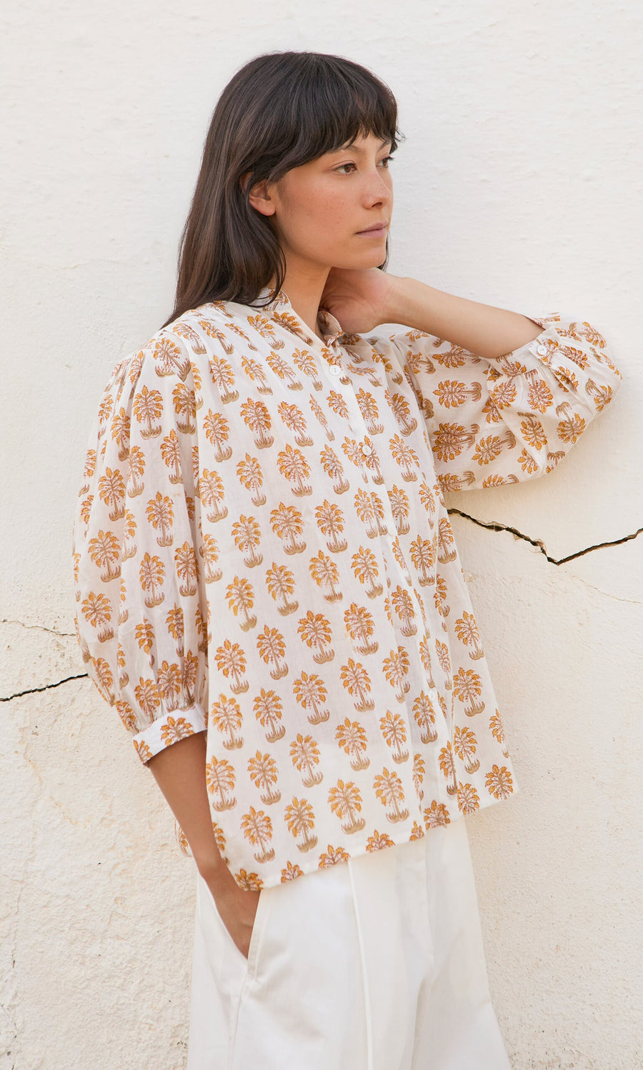 Plümo- Woman in a Palm Shirt and white pants stands against a white textured wall, looking to the side.