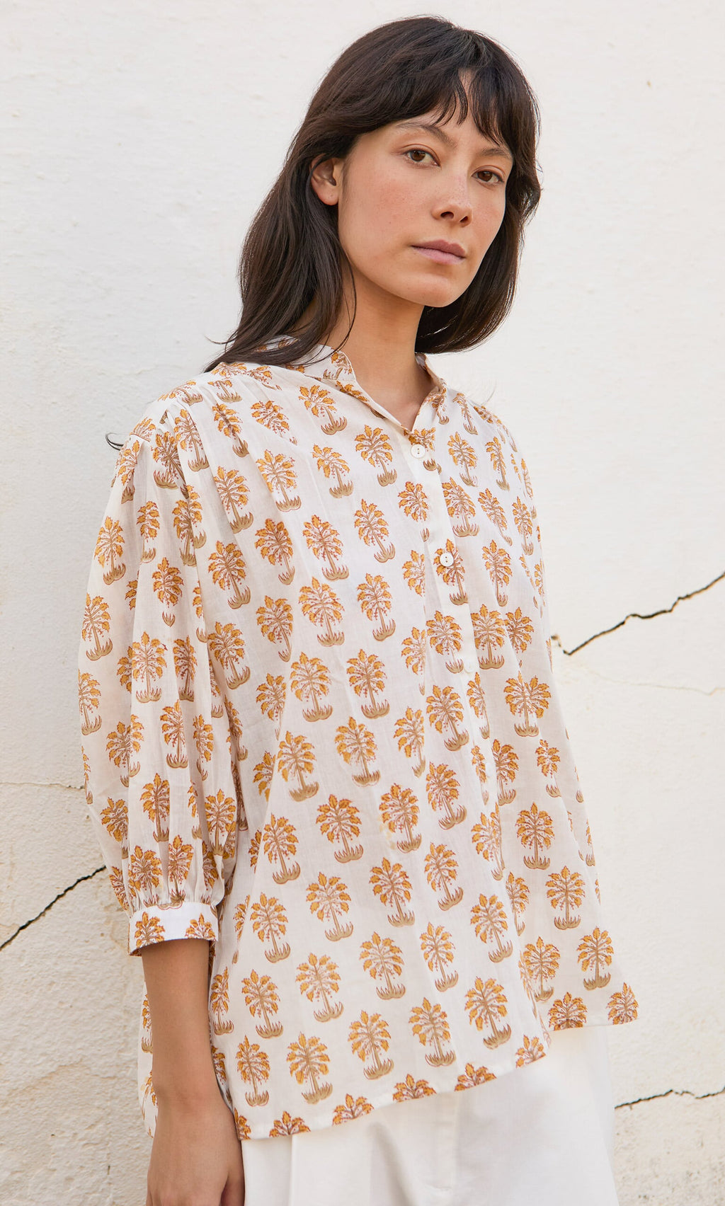 Plümo- Woman wearing the Palm Shirt, a loose blouse with orange floral prints, standing by a white textured wall.