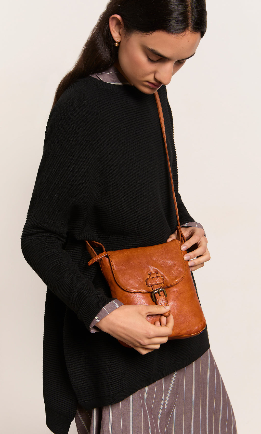 Plümo  Woman in black sweater and striped dress holding the Parcel Bag - Chestnut crossbody, looking down.