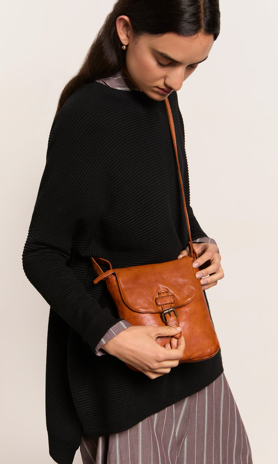 Plümo  Woman in black sweater and striped dress holding the Parcel Bag - Chestnut crossbody, looking down.