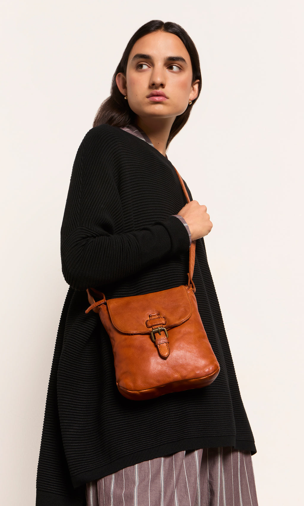 Plümo  Person in black sweater and striped skirt, holding a Parcel Bag - Chestnut, looks to the side.