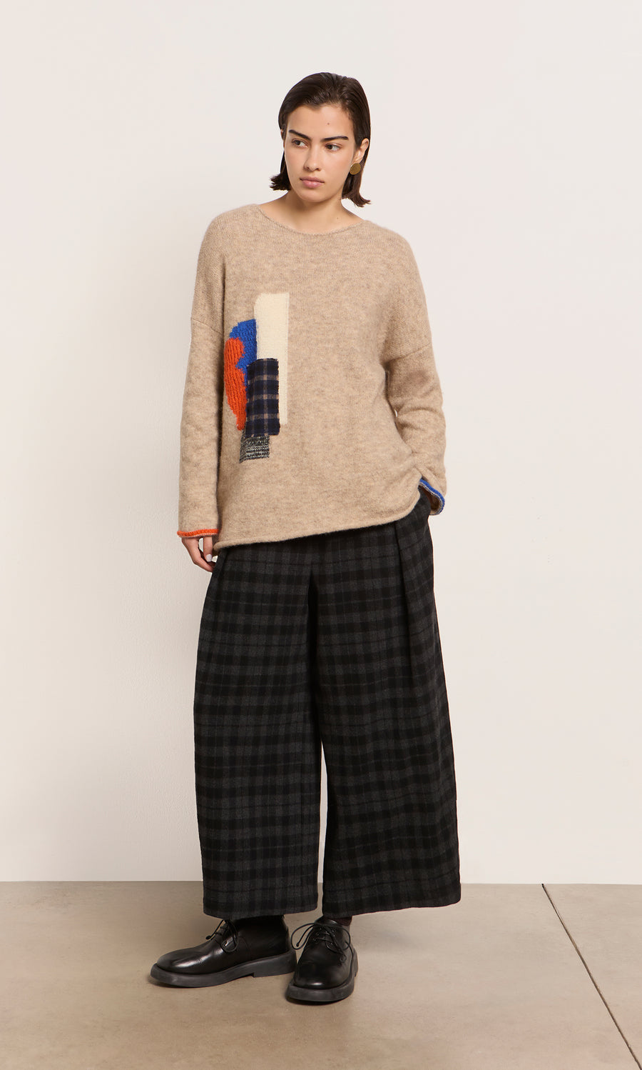 Plümo  Person in a Penrose Jumper with an abstract design and wide, plaid black pants stands against a plain white wall.