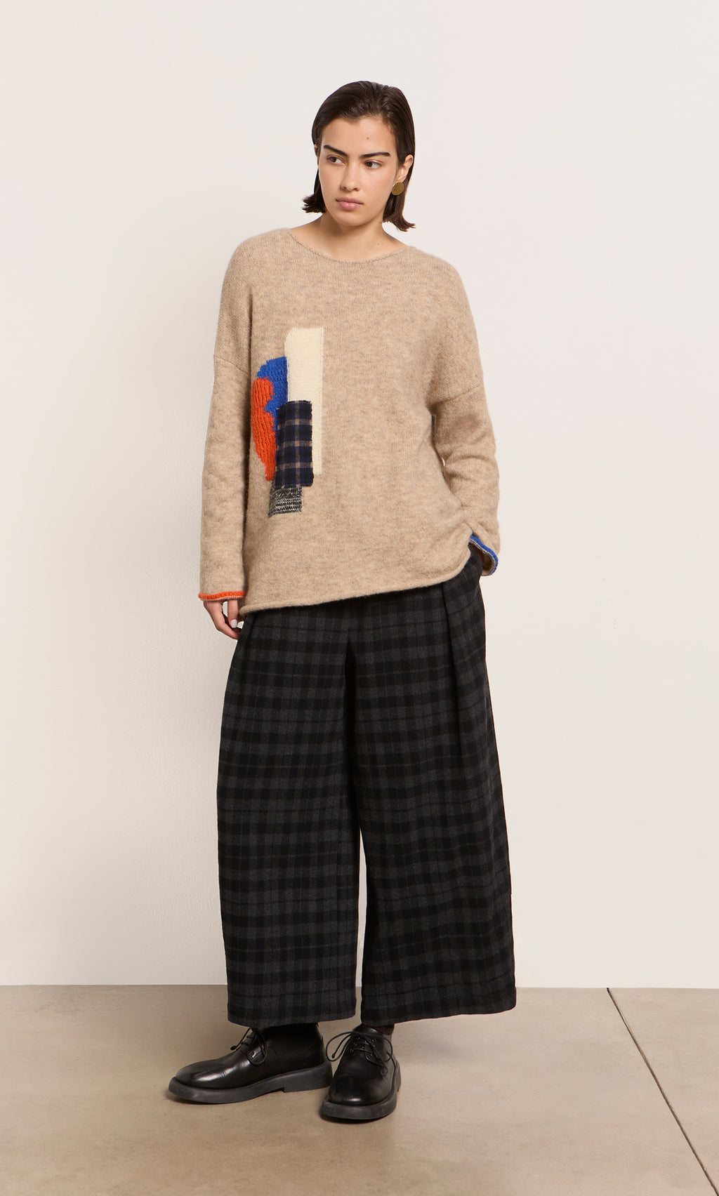 Plümo  Person in a beige sweater and Tartan Flannel Pants made in Italy stands against a white wall.