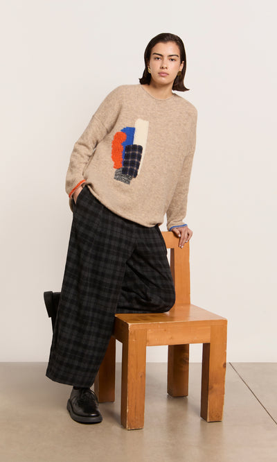 Plümo  Someone in the Penrose Jumper—a lightweight beige abstract knit—leans on a chair, paired with plaid pants and black shoes.