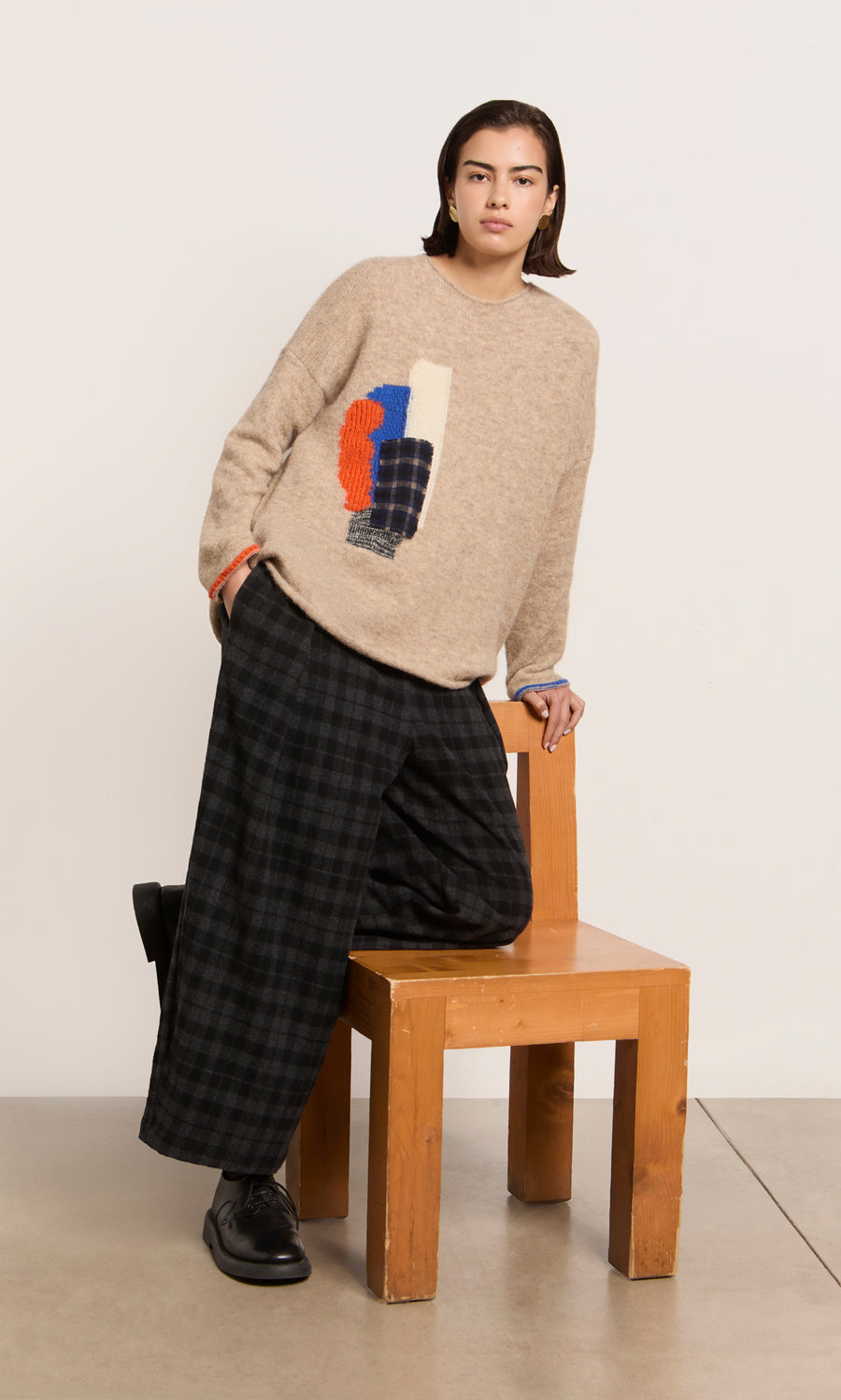 Plümo  Someone in the Penrose Jumper—a lightweight beige abstract knit—leans on a chair, paired with plaid pants and black shoes.
