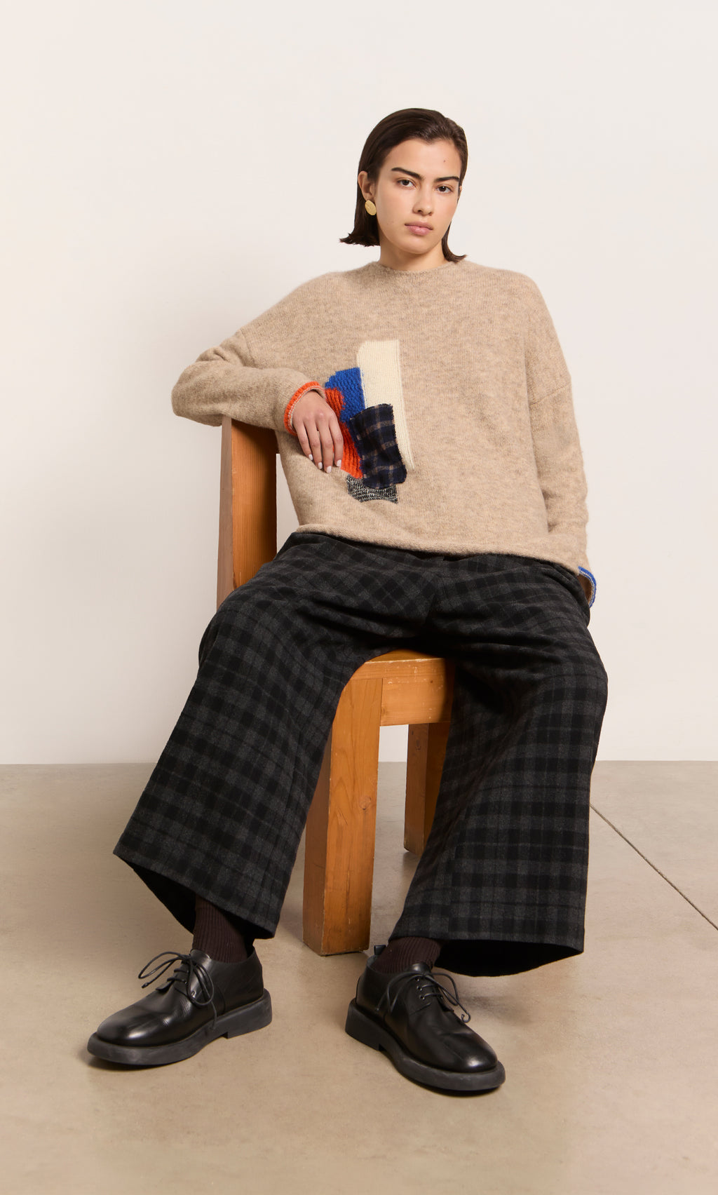Plümo  Person in a beige sweater and Italian Tartan Flannel Pants sits on a wooden chair, looking at the camera.