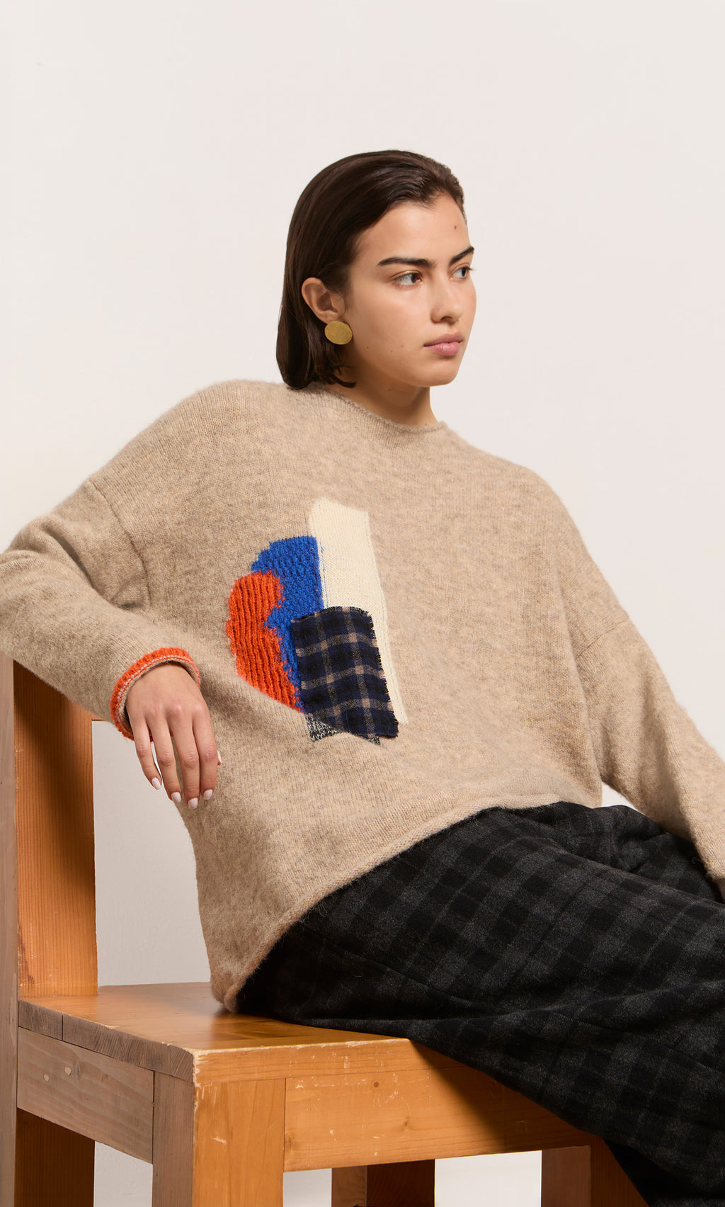 Plümo  Woman in a Penrose Jumper, black plaid pants, and round earrings sits on a wooden chair.