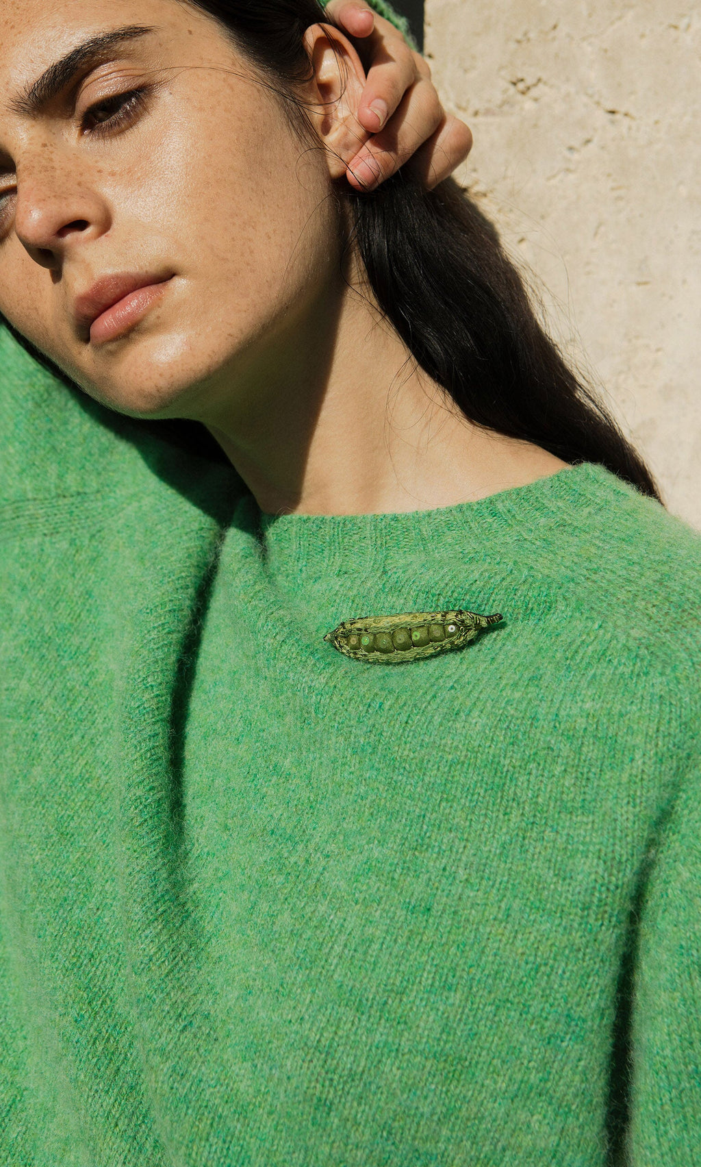 Plümo- Woman in a green sweater wears the Peapod Brooch, standing against a beige wall with arm raised and eyes closed.
