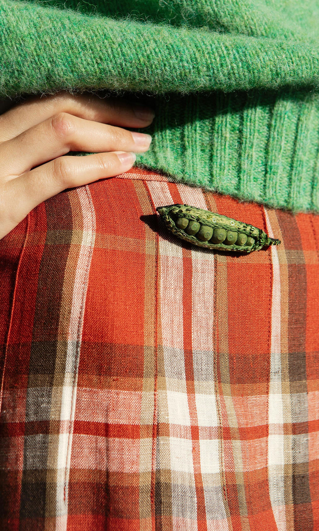 Plümo- A hand rests on a green sweater; an artisan Peapod Brooch is pinned to a plaid skirt.