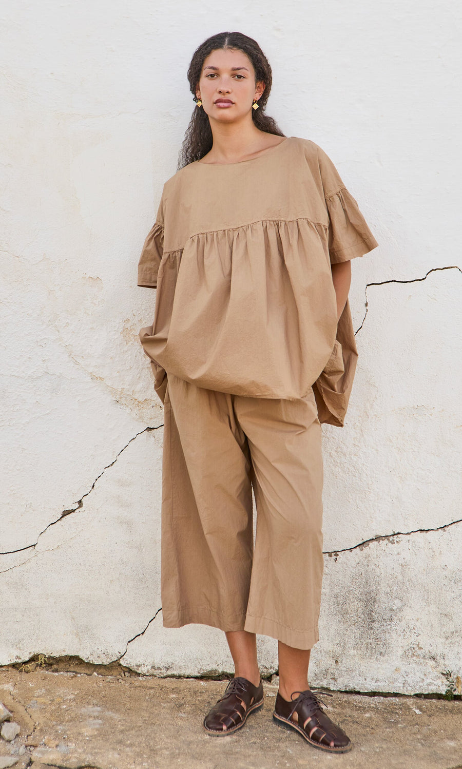 Plümo- Woman standing against a white wall, wearing a loose tan Peplum Top, wide tan pants, and dark brown sandals.
