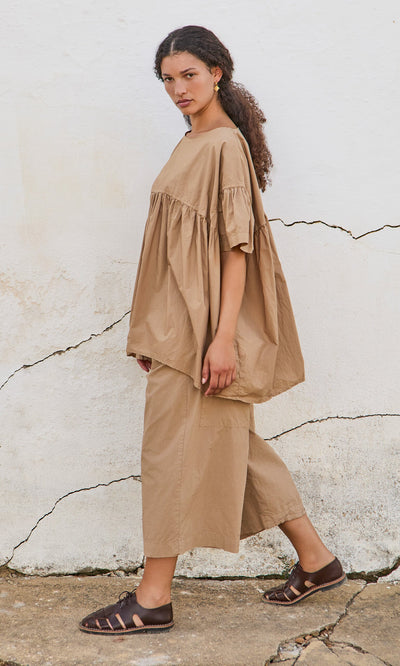 Plümo- Woman in a peplum top and matching wide-leg pants with brown sandals, standing by a cracked wall.