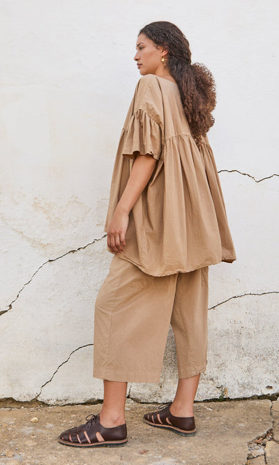 Plümo- A woman in a peplum top and loose tan pants stands sideways against a textured white wall.