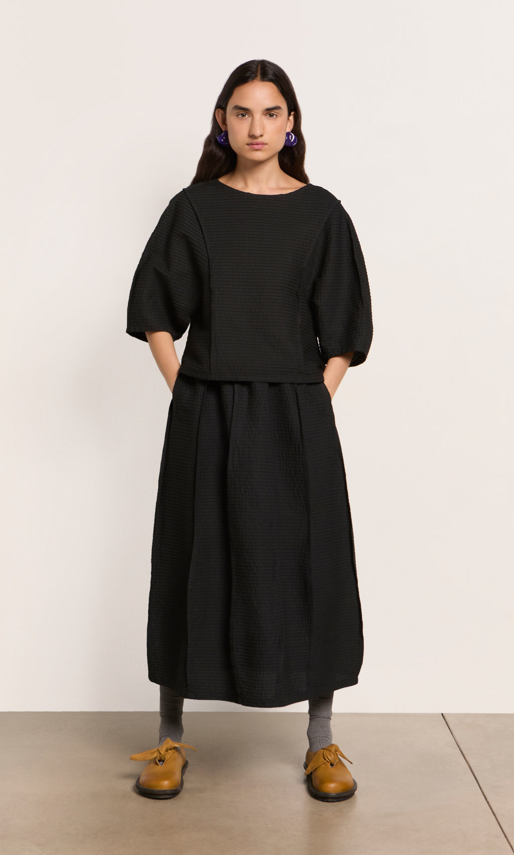 Plümo  Person in a black Poko Top cotton blouse and skirt, gray socks, and tan shoes stands against a plain background.