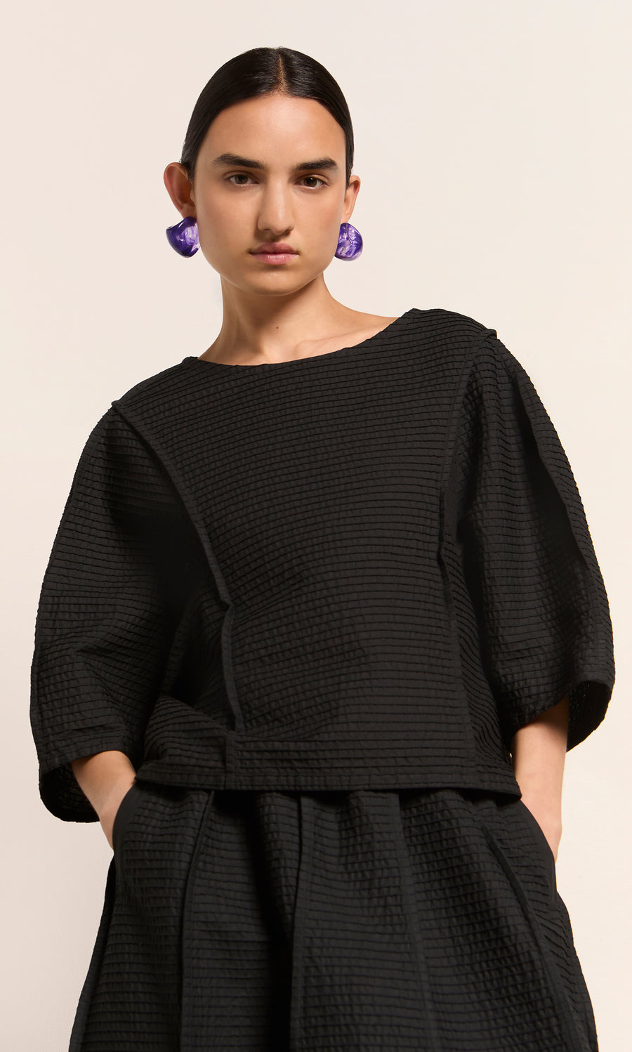Plümo  Woman in a textured black Poko Top with wide sleeves and purple earrings, against a light background.