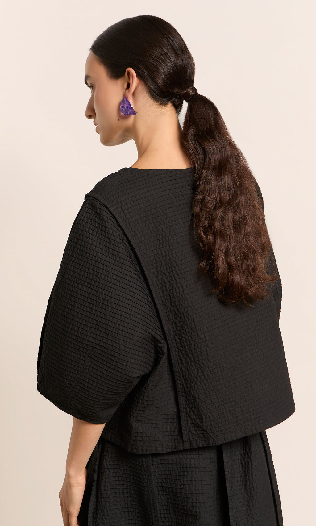 Plümo  Woman with long brown hair in a low ponytail, wearing the Poko Top and purple earrings, facing sideways.