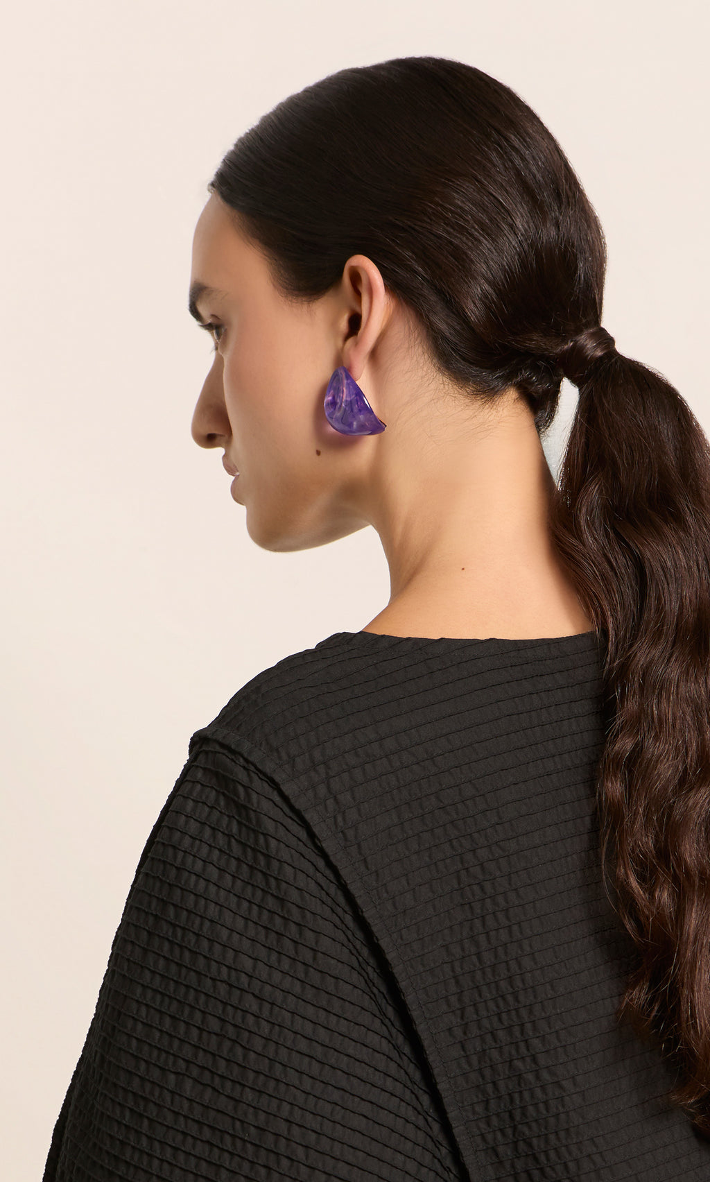 Plümo  Woman with dark hair in a low ponytail, wearing Viola Earrings and a black textured top, facing left.