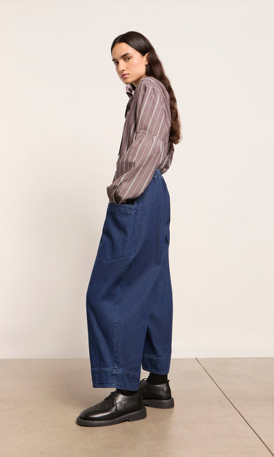 Plümo  Person in a striped brown shirt, Polder Pants - Original Denim, and black shoes stands sideways on a beige floor.