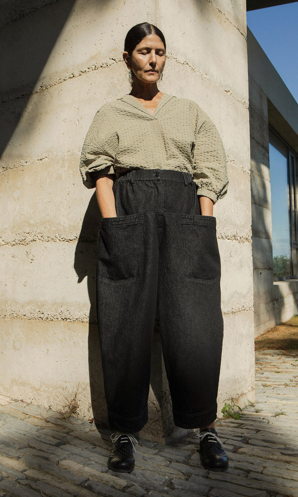 Plümo- A woman stands outside by a concrete wall in a beige top and Polder Pants - Washed Black, hands in her pockets.