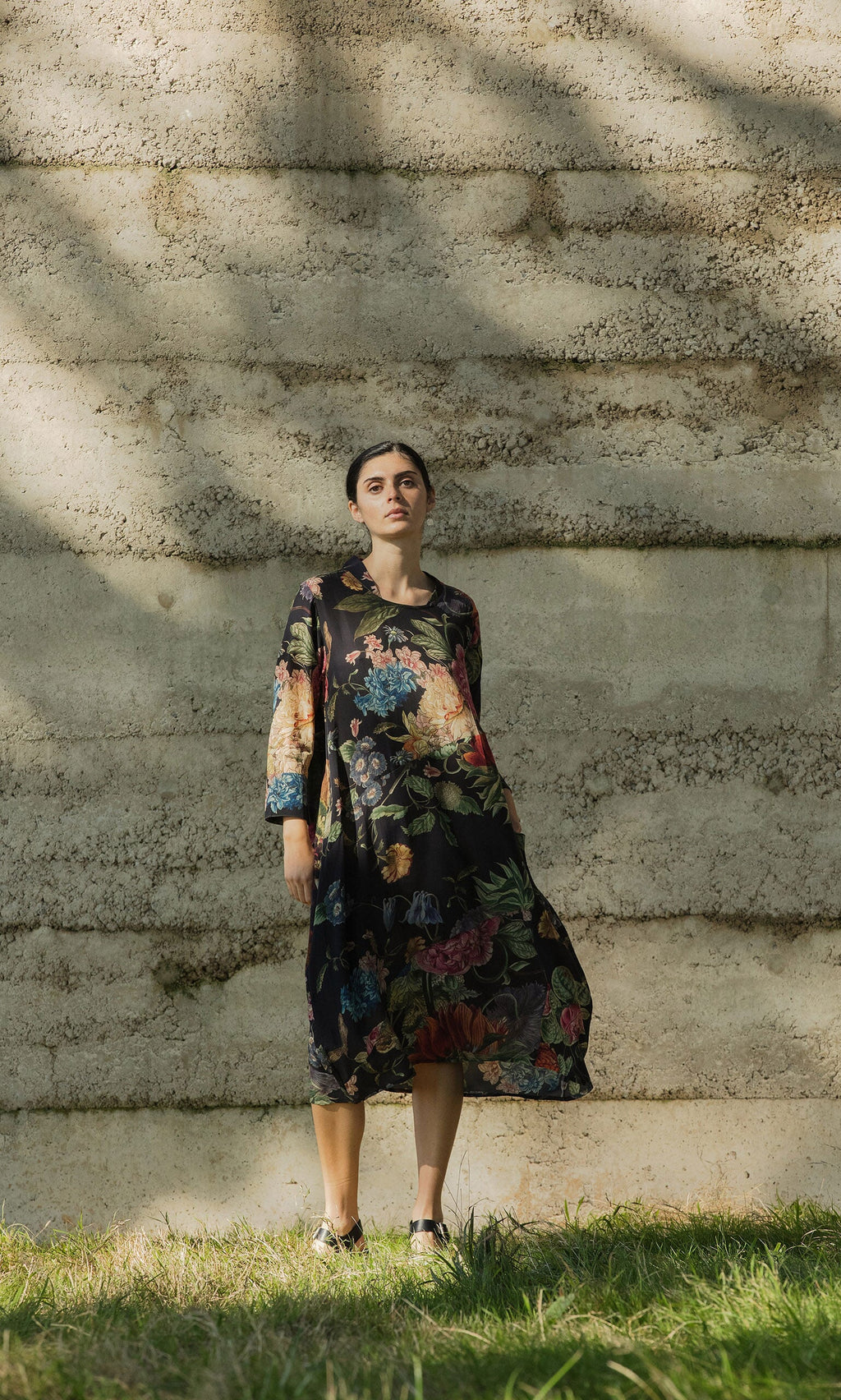 Plümo Woman in the Primula Dress stands on grass by a stone wall, looking at the camera.