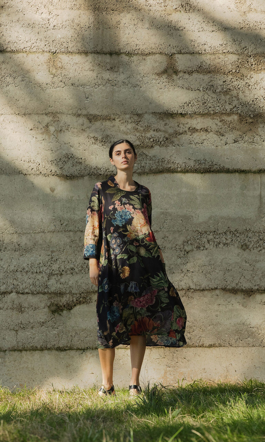 Plümo Woman in the Primula Dress stands on grass by a stone wall, looking at the camera.