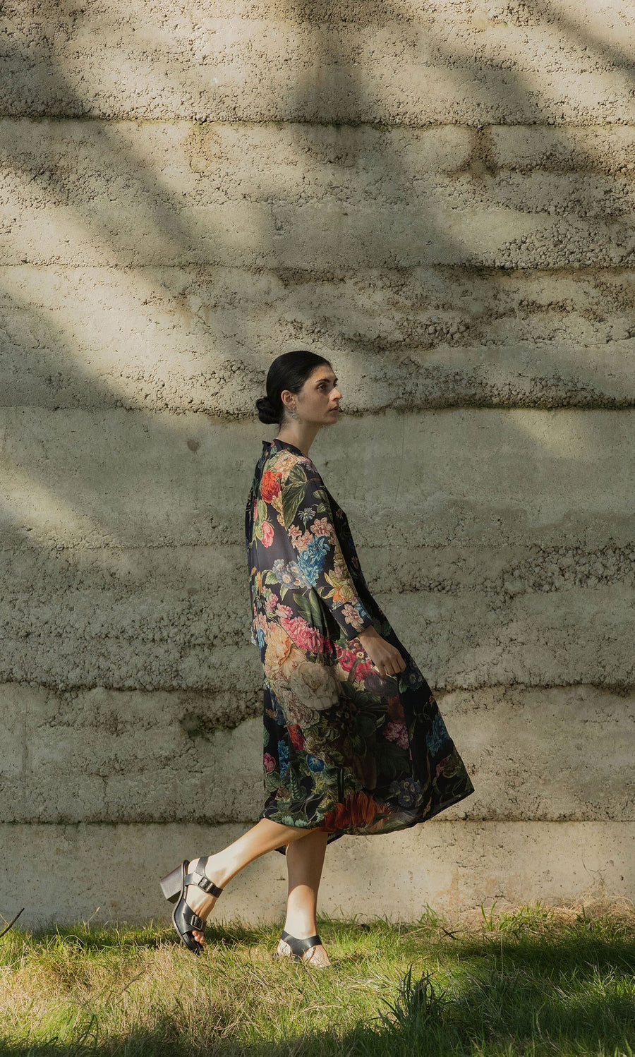 Plümo - Woman in a Primula Dress and sandals walks on grass beside a textured concrete wall with shadow patterns.