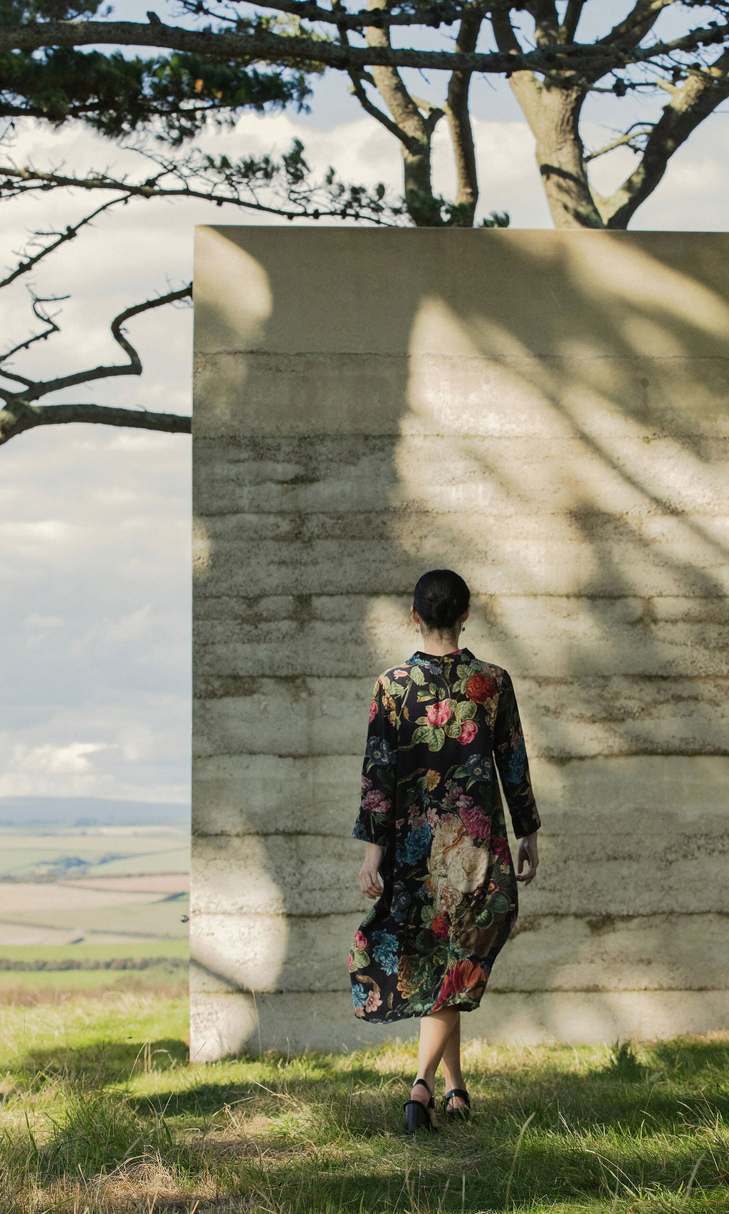 Plümo - A person in a Primula Dress walks toward a concrete wall outdoors, surrounded by trees and open landscape.