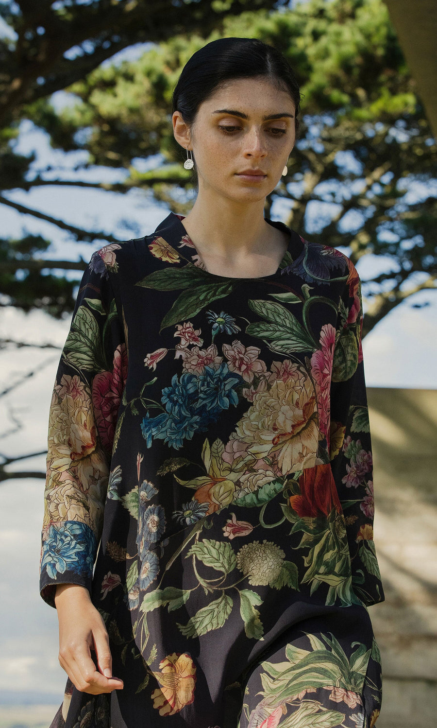 Plümo Woman wearing Primula Dress stands outdoors with trees and a blue sky in the background.
