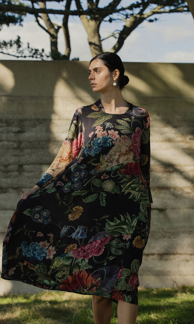Plümo - Woman in a Primula Dress stands outdoors on grass, sunlight and tree shadows in the background.