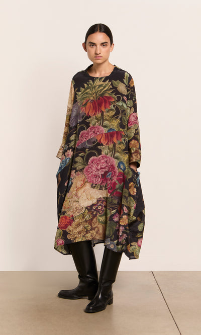 Plümo  Woman wears the Primula Dress—a loose, floral A-line—paired with black knee-high boots, standing by a plain wall.