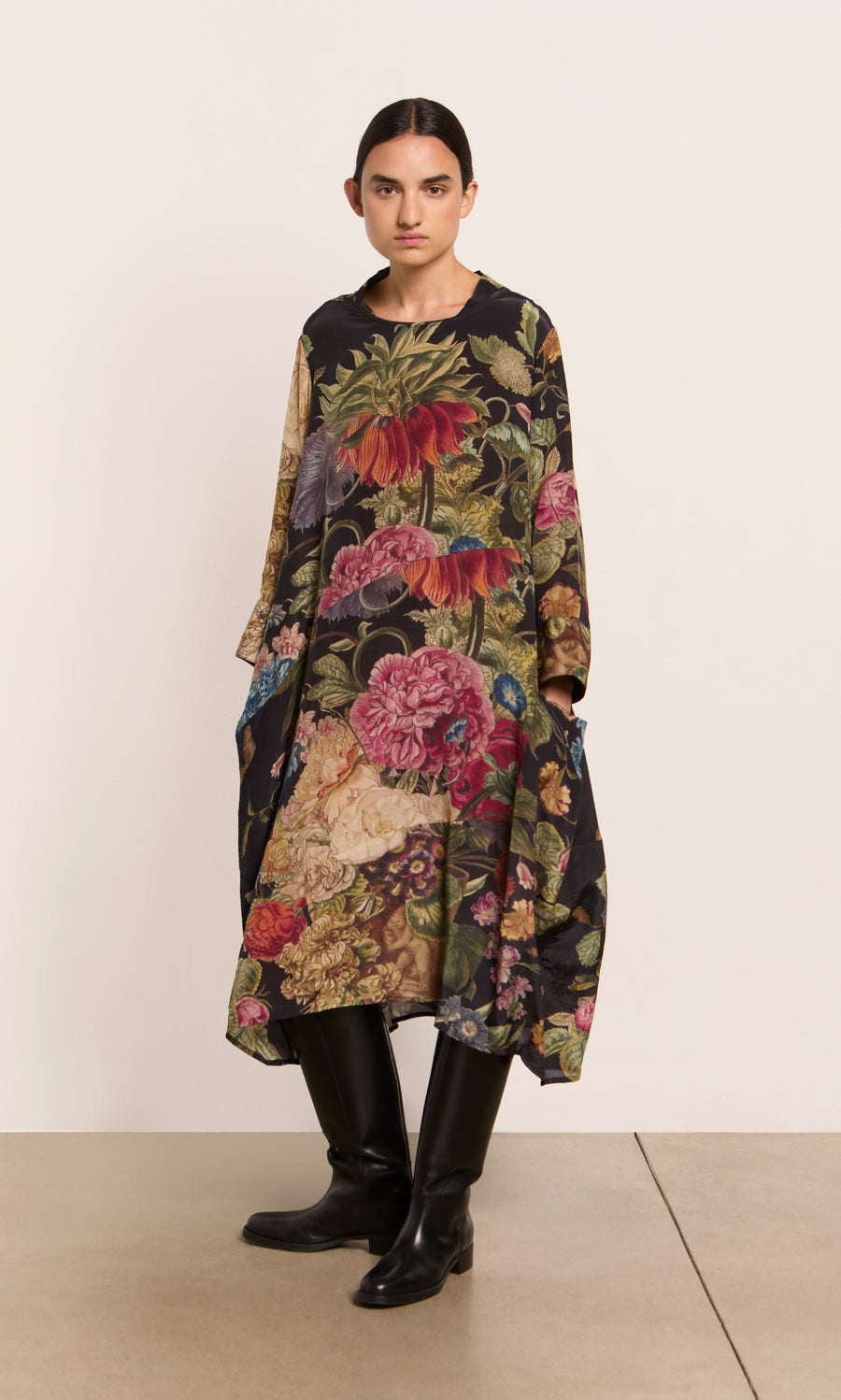 Plümo  Woman wears the Primula Dress—a loose, floral A-line—paired with black knee-high boots, standing by a plain wall.