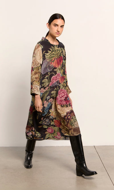 Plümo  Person in the Primula Dress with asymmetric pockets and black boots walks on a light floor near a plain wall.