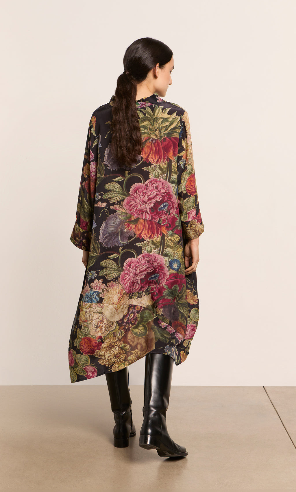 Plümo  Woman in the Primula Dress and black boots, back to camera, standing against a plain wall.