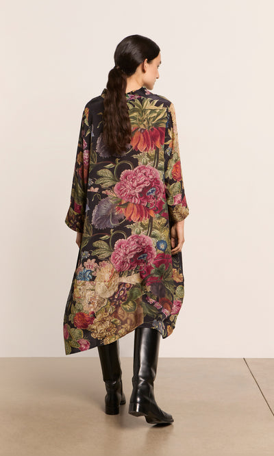Plümo  Woman in the Primula Dress and black boots, back to camera, standing against a plain wall.
