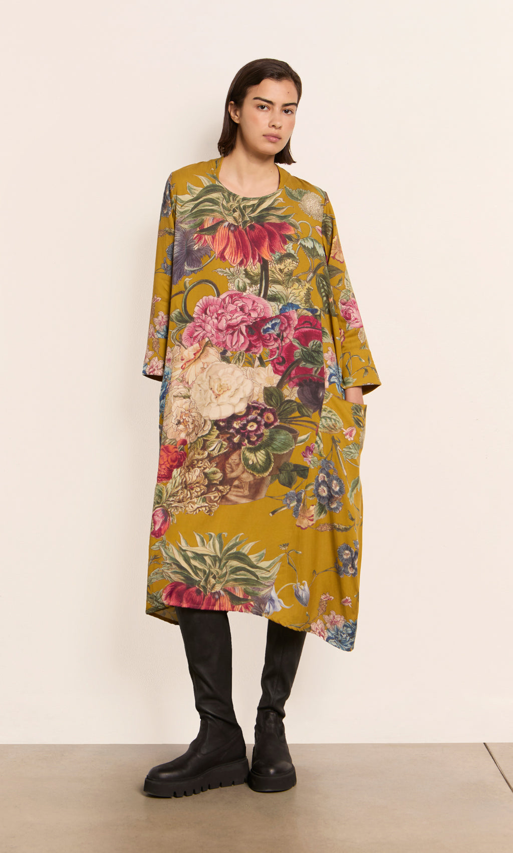 Plümo  Woman wears the Primula Dress - Gold with black boots, standing against a plain off-white wall.