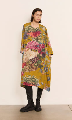 Plümo  Woman wears the Primula Dress - Gold with black boots, standing against a plain off-white wall.