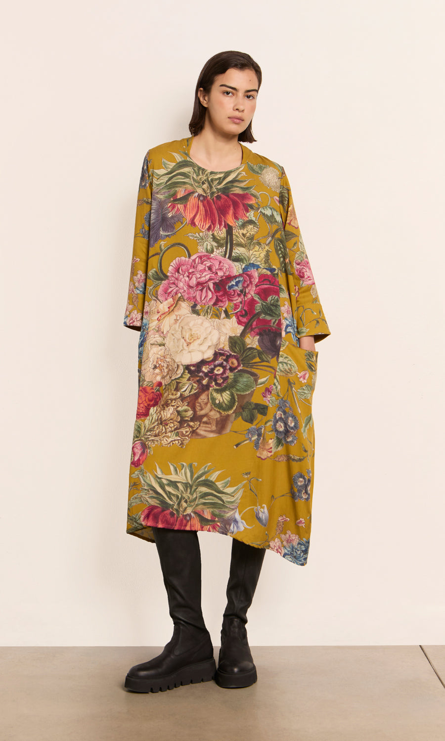 Plümo  Woman wears the Primula Dress - Gold with black boots, standing against a plain off-white wall.