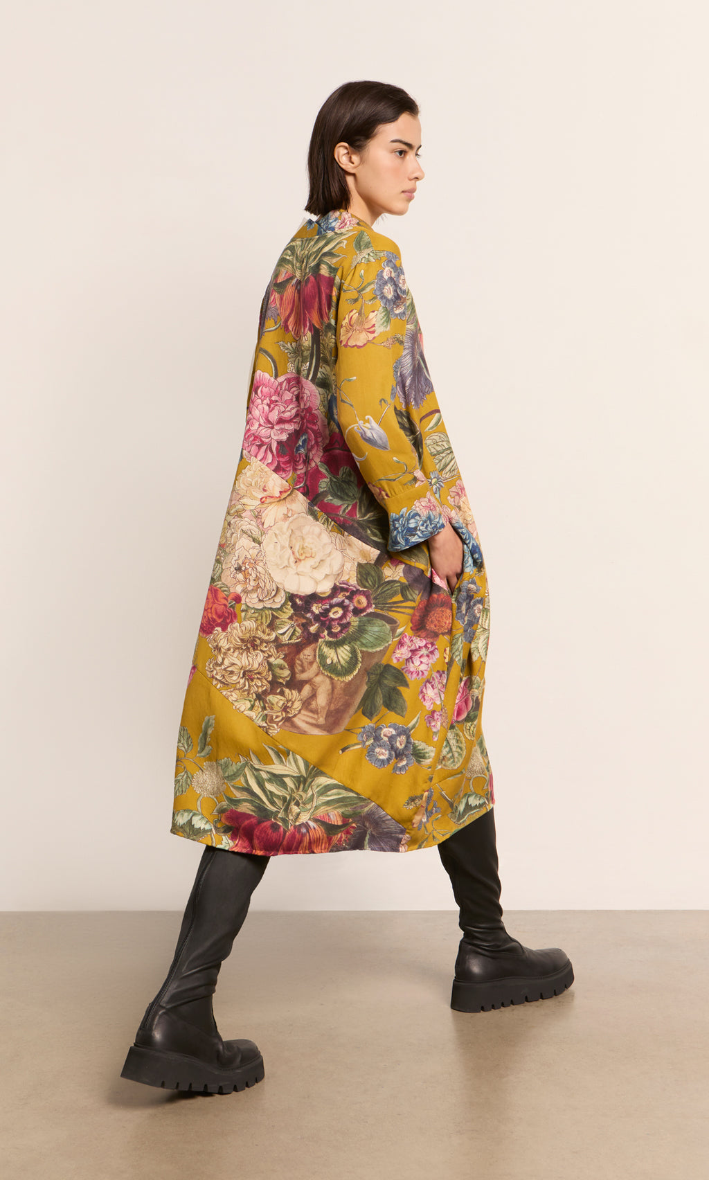 Plümo  Woman in the Primula Dress - Gold, walking sideways indoors in a relaxed fit with chunky boots.