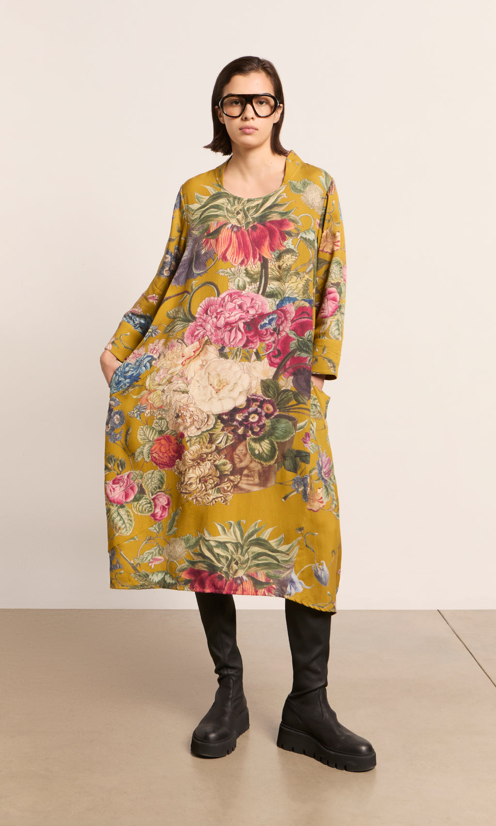 Plümo  Woman in glasses wears the Primula Dress - Gold and black boots, standing indoors.