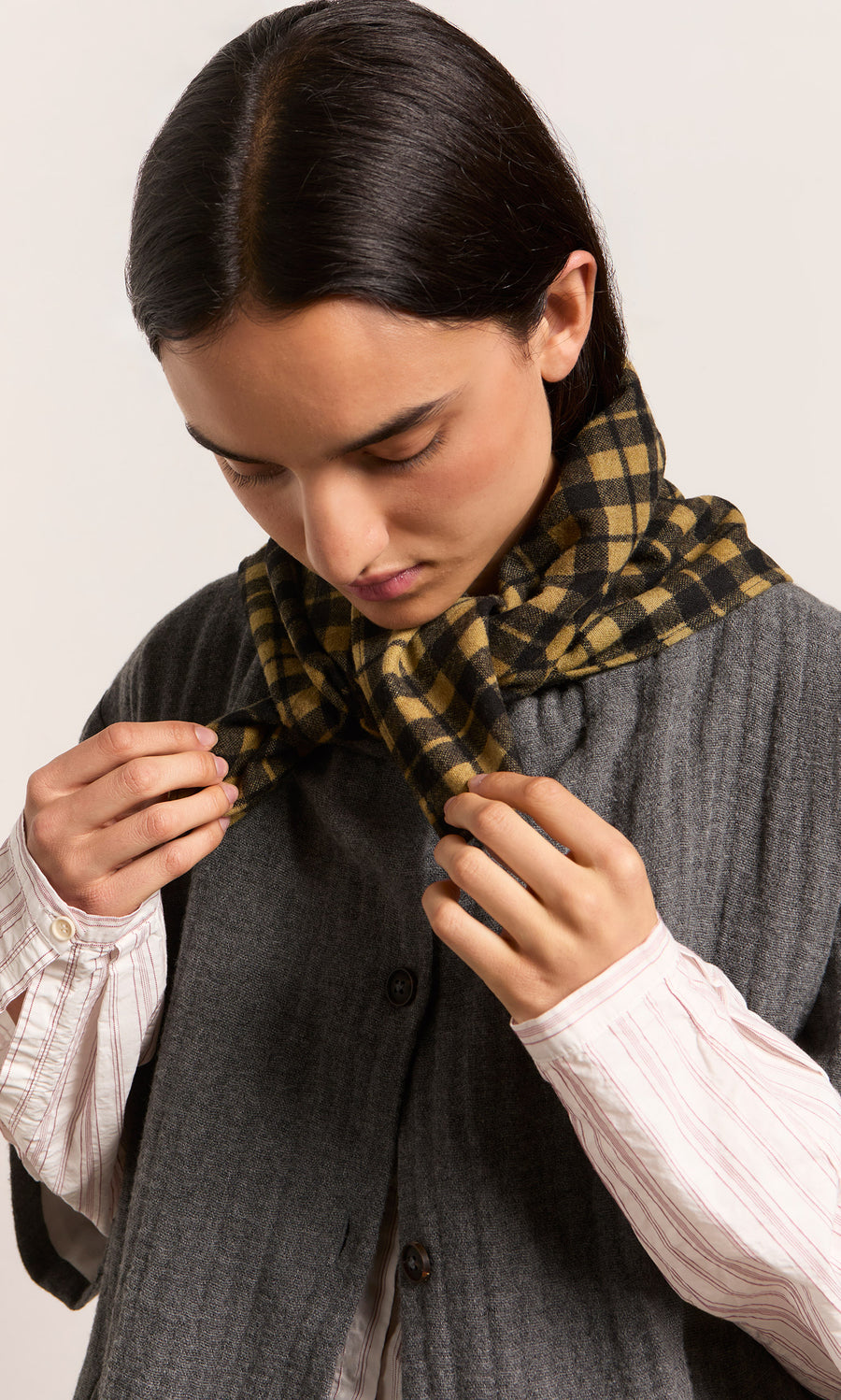 Plümo  Person looking down, tying a Quadrant Necktie over a gray buttoned vest and striped shirt.