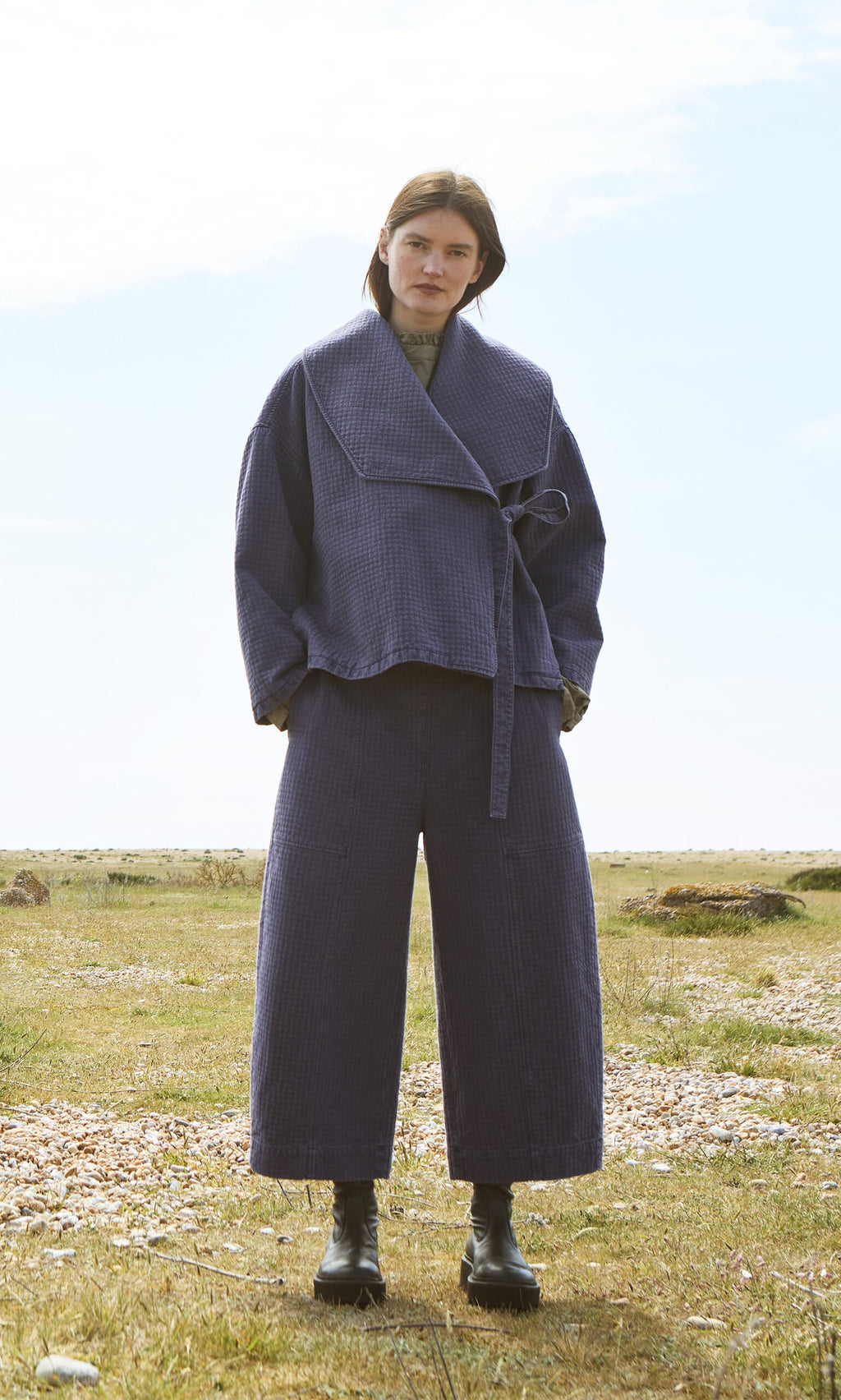 Plümo- Woman outdoors in Calow Pants—checked curved-leg trousers—with a blue jacket, black boots; made in London.