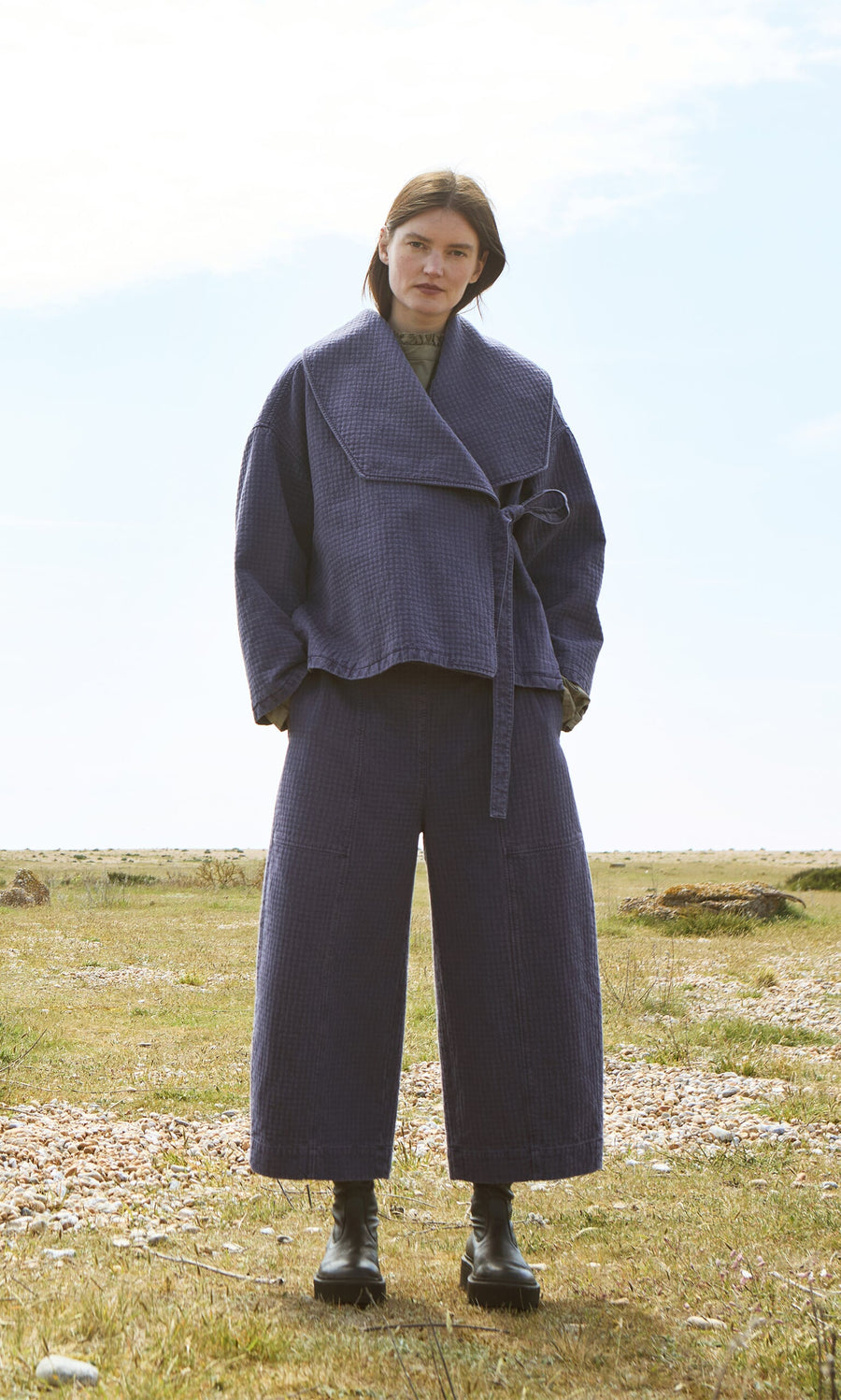Plümo- Woman outdoors in Calow Pants—checked curved-leg trousers—with a blue jacket, black boots; made in London.