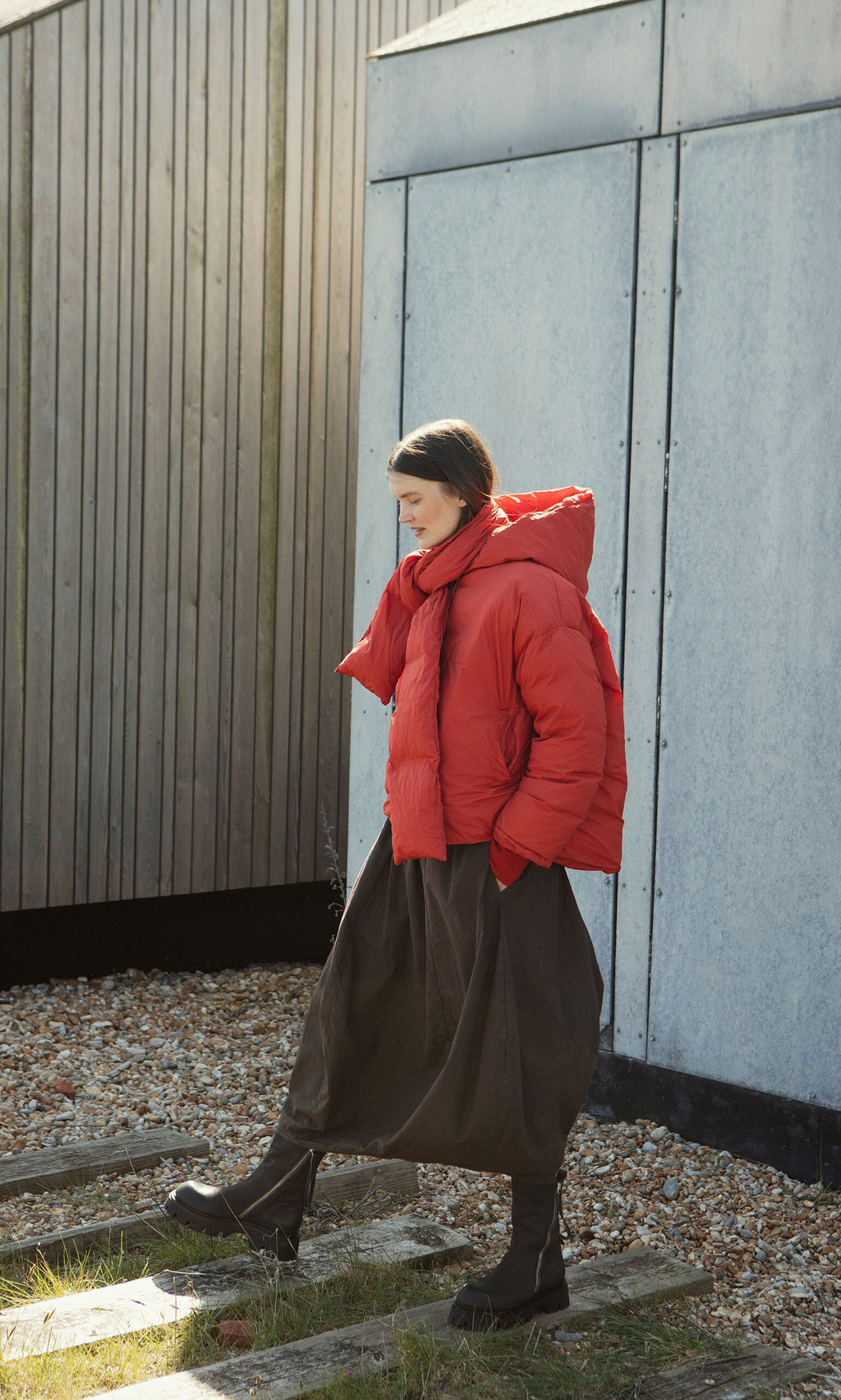 Plümo- Woman in a Red Puffer coat stands outside by a wooden and metal building, wearing a long dark skirt.